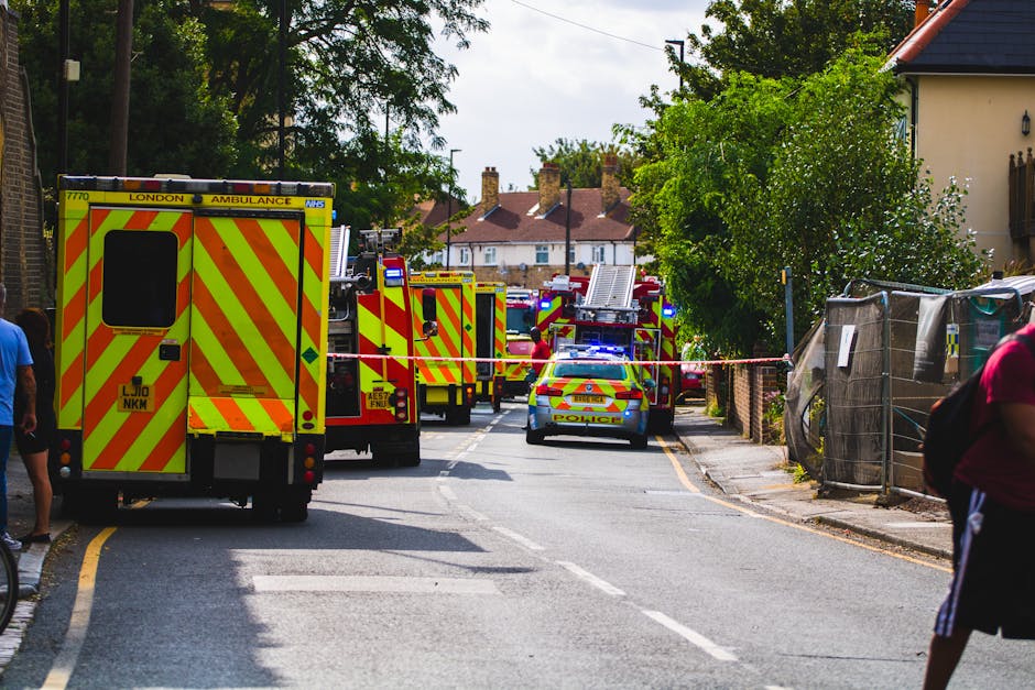 A street scene showing multiple emergency response vehicles, including a London ambulance and several fire trucks with bright yellow and red chevron markings, parked along a residential area with tree