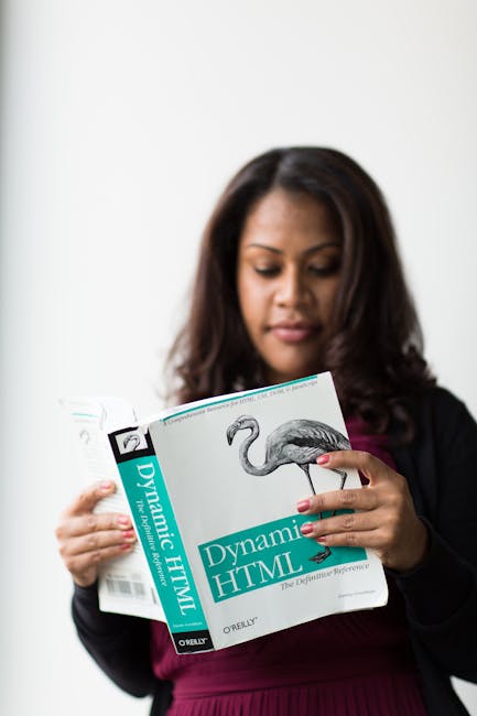 A woman with dark hair, dressed in a black blazer and maroon top, is holding and reading a book titled 'Dynamic HTML' with a cover illustration of a flamingo. The background is plain white, and her fo